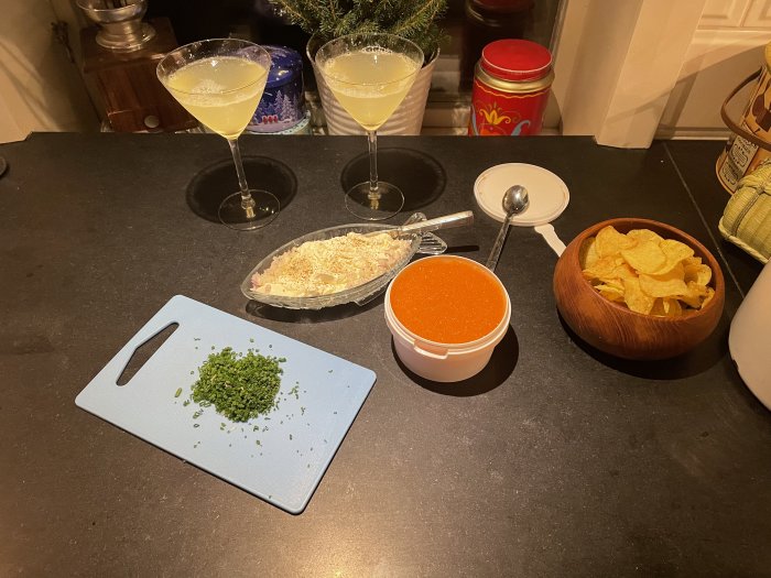 Matlagningsscenario med hackade grönsaker, dippsås, chips, cocktailglas och riven ost på köksbänk.