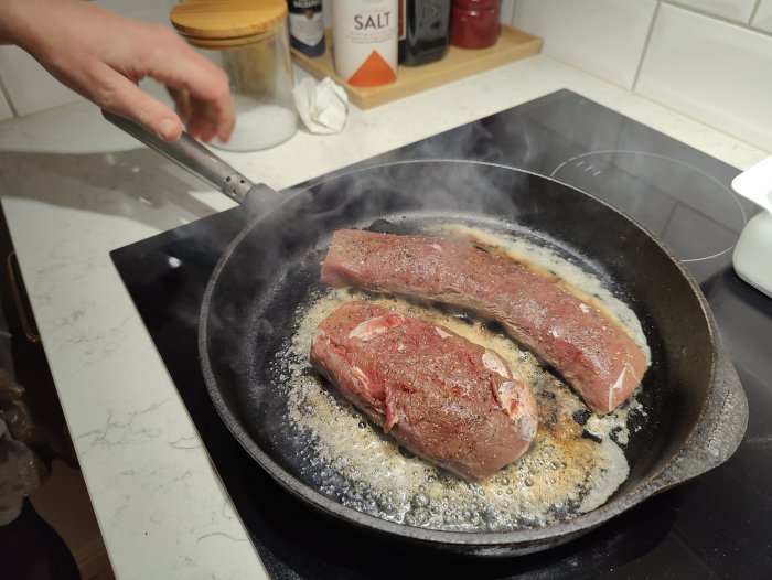 Två stekta biffar i en stekpanna, hand som vänder kött, smält smör, kryddor i bakgrunden.