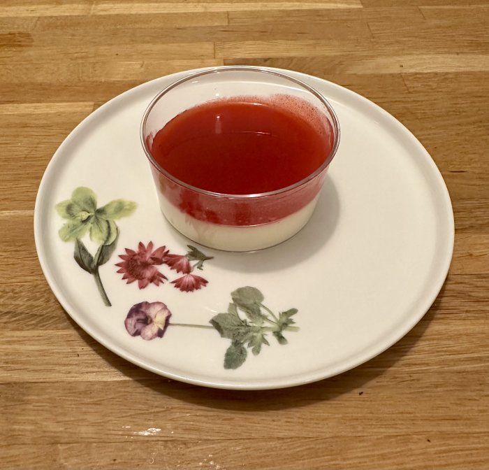 Ett glas med dessert med röd gelé överst, på en blommigt dekorerad tallrik, mot träbakgrund.