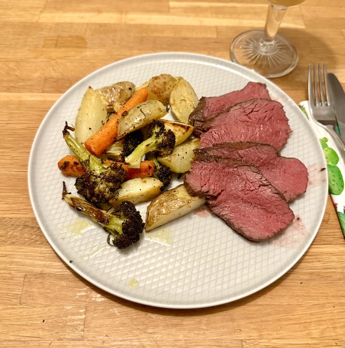 En tallrik med skivor av medium-rare stekt kött och rostade grönsaker. Bakgrund av träbord, gaffel, tomt glas.