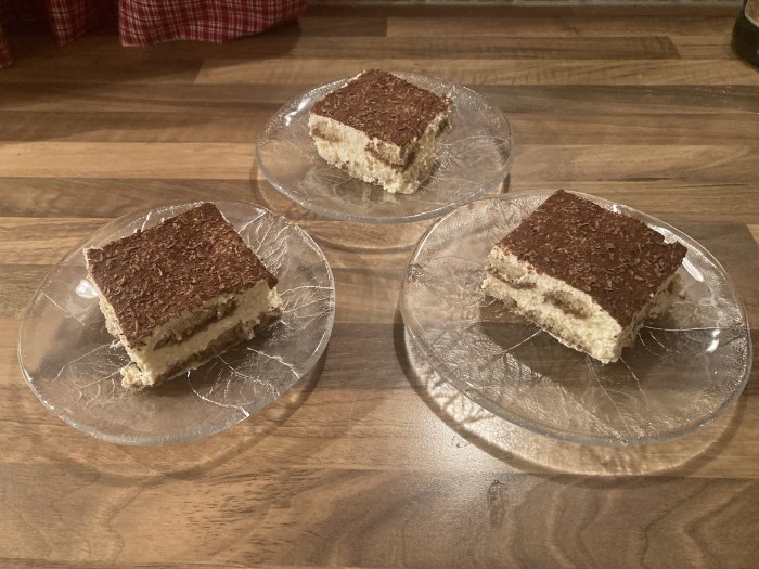 Tre bitar av tiramisu på glastallrikar, på ett träbord. Dessert, bakverk, kaffe, kakaoströssel, hemlagat eller café.