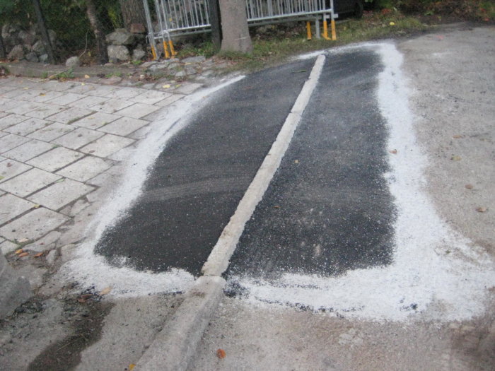 Vägarbete med nylagt asfalt och vit vägmarkering vid kantsten. Trottoar och grus syns också.