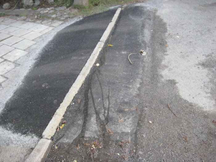 En lagad asfaltsyta bredvid en trottoarkant, med löv och smuts, troligen efter vägarbete.