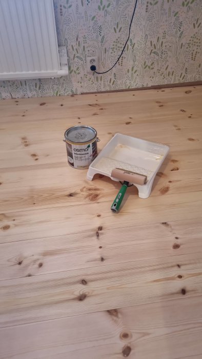 Renovering inomhus, trägolv, färgburk, rollerfat, vägg med tapet, eluttag, vit radiator.