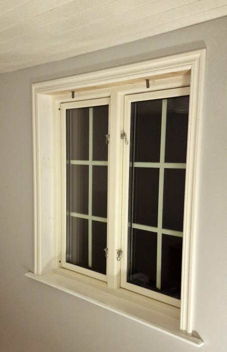 Vitt snedtakfönster med korsformade spröjs, öppna handtag och mörk utsikt.