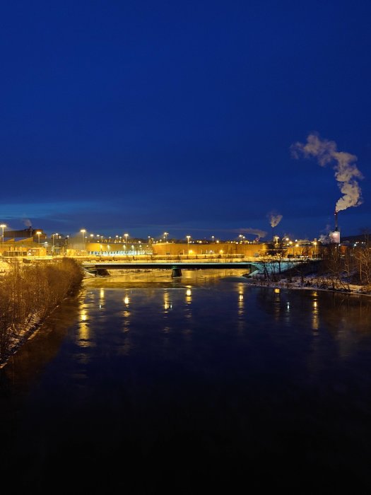 Kvällsstadssiluett med flod, bro, industrianläggning och rök från skorstenar mot blå himmel.
