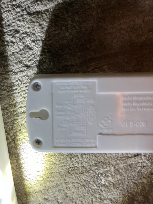 Närbild av en etikett på elektronisk utrustning med text och ledningsdiagram, upplyst av starkt ljus, mot texturerad bakgrund.