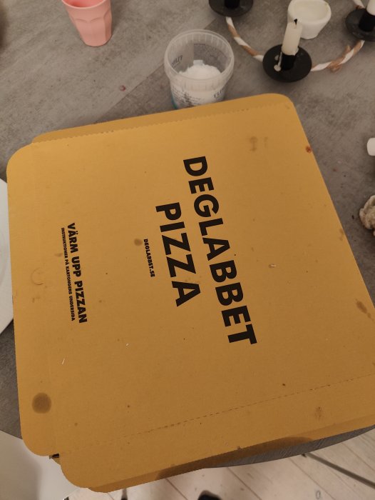 Gul pizzakartong märkt "DEGLABBET PIZZA", på ett bord med ljusstakar och vitt pulver.