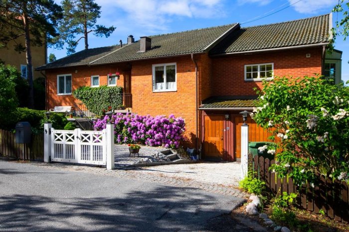 Tvåvånings tegelhus med garage, vit grind, grönska, blå himmel, och blommande buskar.