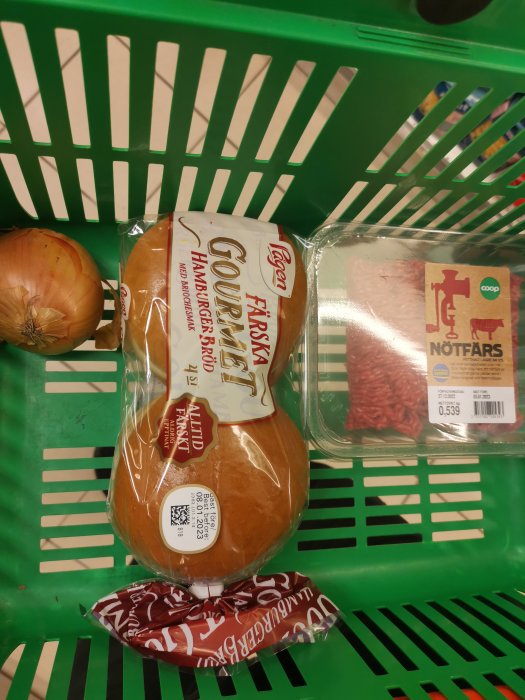 Grönt kundvagnskorg med hamburgerbröd, nötfärs och lök. Matinköp.