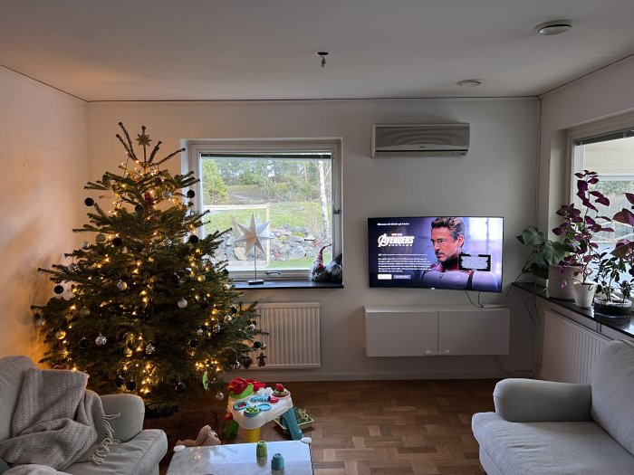 Vardagsrum, julgran med ljus, TV med film, soffa, barnleksaker, fönster med utsikt, växt på fönsterbräda.