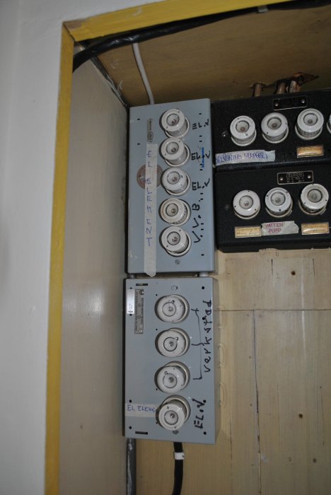 Äldre säkringsskåp med skruvsäkringar, etiketter, på en vägg med träpanel och kabelrör.