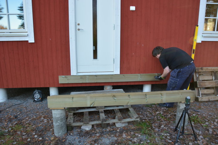 Person bygger trästaket vid röd stuga, använder verktyg, nivåmätare, trädgård, grusmark.