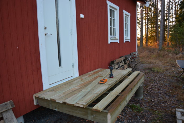 Nybyggd trädäck vid röd stuga, skruvdragare på plank, skog i bakgrunden, skymning eller gryning.