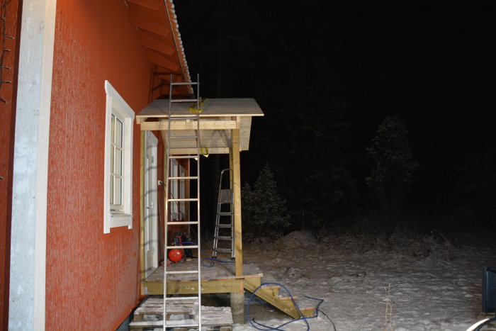 Ett rött hus med veranda under konstruktion om natten, omgiven av snö och skog.