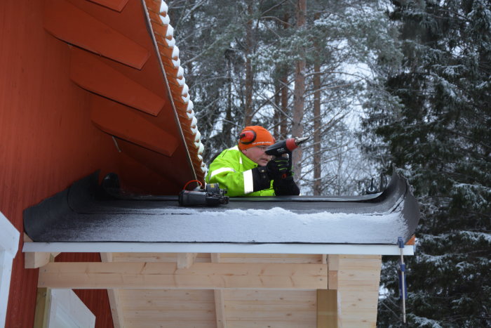 Arbetare med hörselskydd och reflexjacka använder borr på snötäckt takkant framför skog.