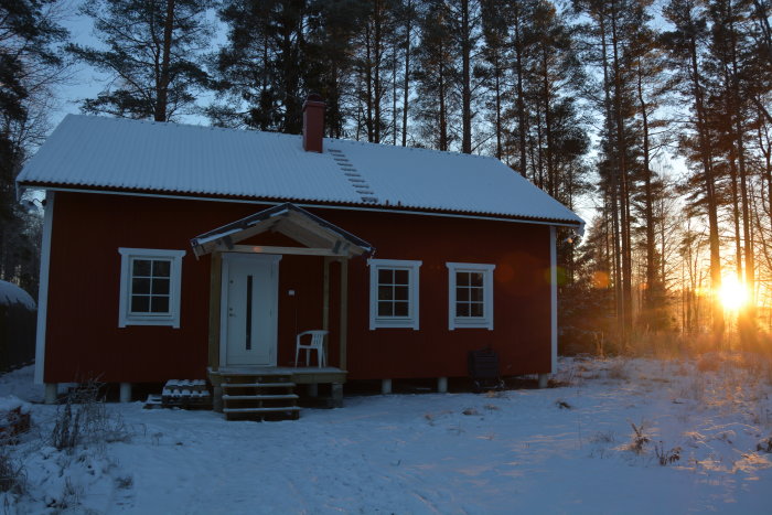 Rött hus i snöigt landskap med skogsbakgrund vid solnedgång.