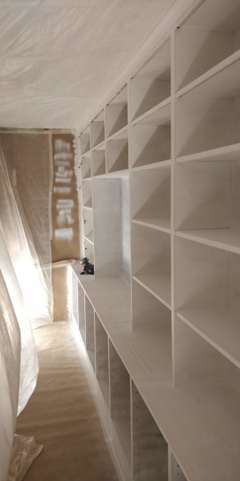 Vitmålad inbyggd bokhylla i ett utrymme under renovering, täckt med plastskydd och arbetsmaterial synligt.
