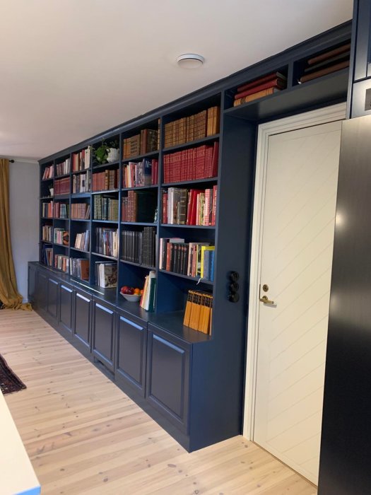 Mörkblå bokhylla med böcker, integrerad vit dörr och trägolv.