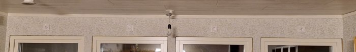 Inomhus, tapetvägg, vita dörrkarmar, fönster, elektriska uttag, hängande glödlampa, taklampa, träbjälkar.