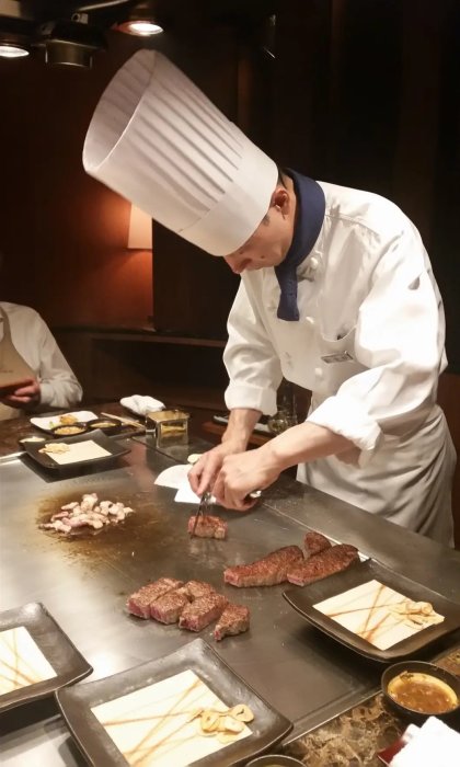 Kock i hög hatt tillagar kött på teppanyakigrill i restaurangmiljö.
