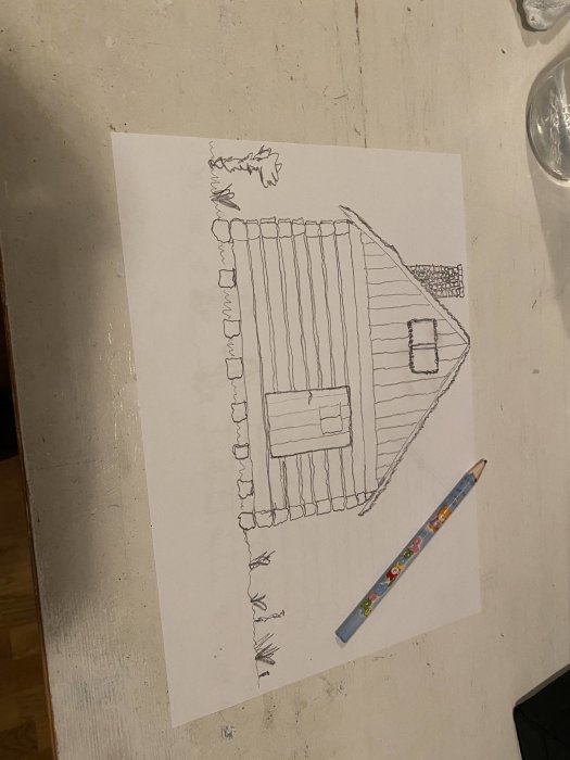 Teckning av ett hus, träd, gräs och staket på ett vitt papper med en blyertspenna bredvid.