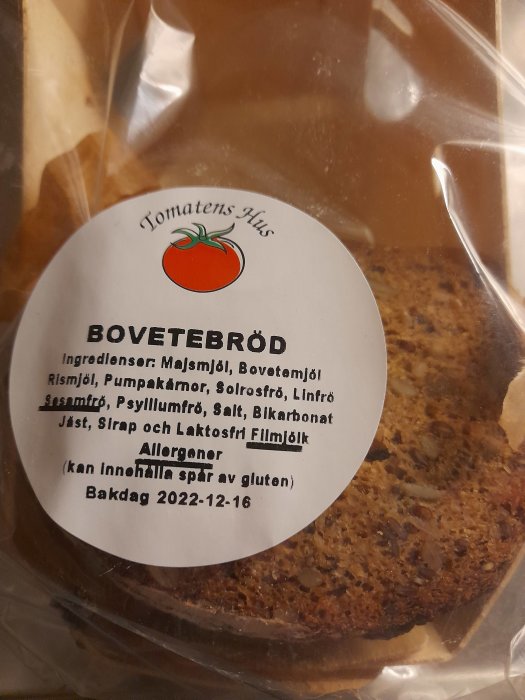 Etikett på bovetbröd med ingredienser, allergeninformation, bakdatum. Synlig skiva bröd bakom. Transparent plastförpackning.