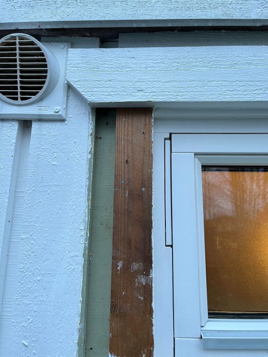 Del av en byggnad med ventilation, fönster och avskalad målning på träelement.