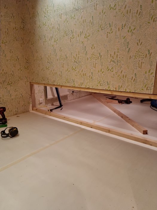 Träkonstruktion vid vägg, mönstrad tapet, verktyg och hörselskydd på golvet, inomhus renoveringsarbete pågår.