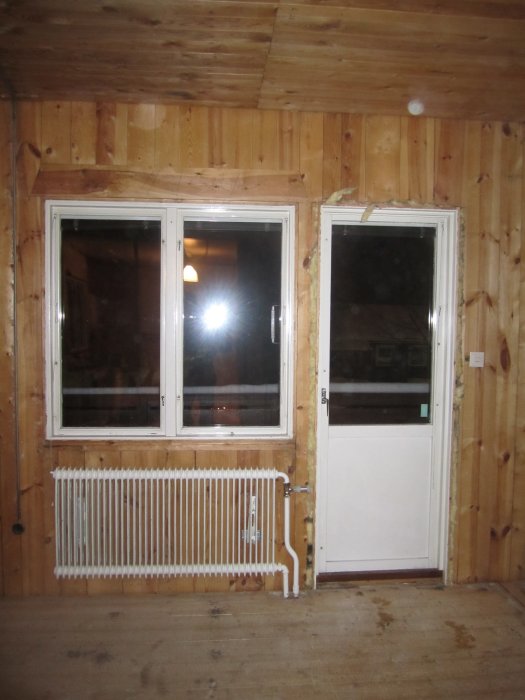 Puuverhoiltu huoneen seinä, jossa on ikkunat ja ovi, alla valkoinen patteri. Taustalla kodikkuutta luova lämmin valaistus.