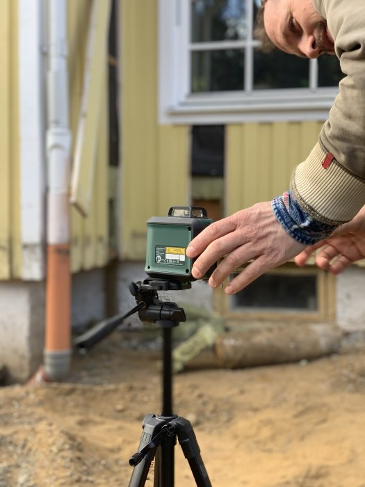 Person justerar nivelleringsinstrument på stativ framför hus, fokus på händer och verktyget, suddig bakgrund.