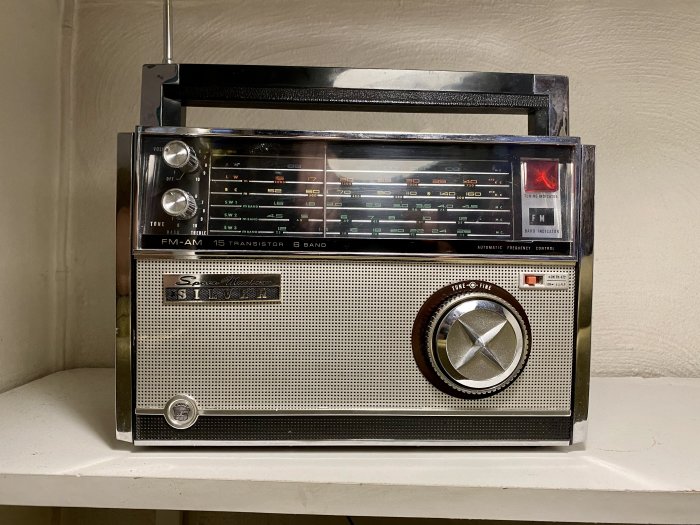 En vintage Silver-tonad transistorradio med FM/AM, vridreglage och utdragbar antenn, placerad på en hylla.