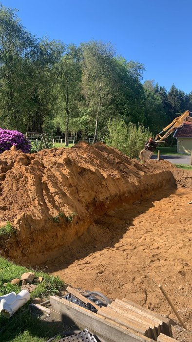 Grävmaskin vid arbete, stort sandberg, grönska i bakgrunden, blå himmel, byggarbetsplats.