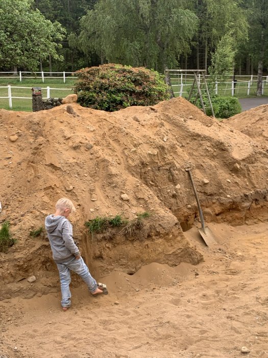 Barn klättrar på sandhög nära skottkärra och spade, grönska och staket i bakgrunden.