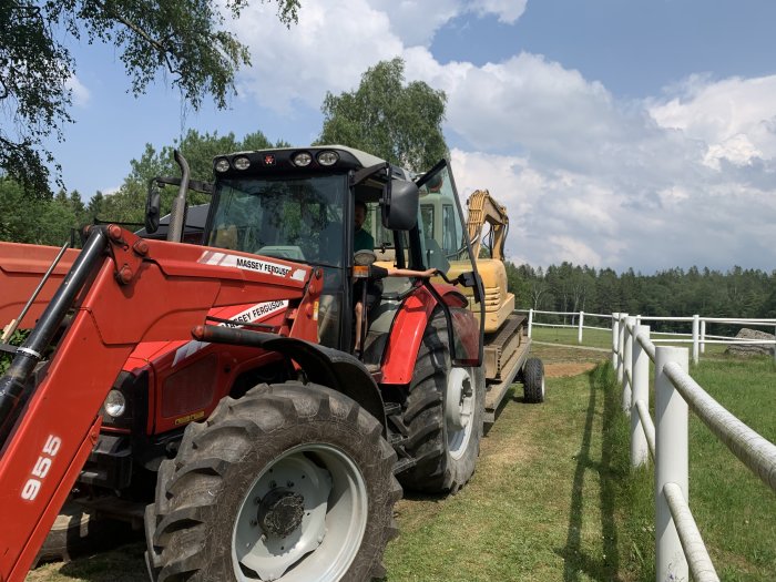 Röd Massey Ferguson traktor med frontlastare, person i hytten, transporterar gul grävmaskin på släp, lantlig miljö.