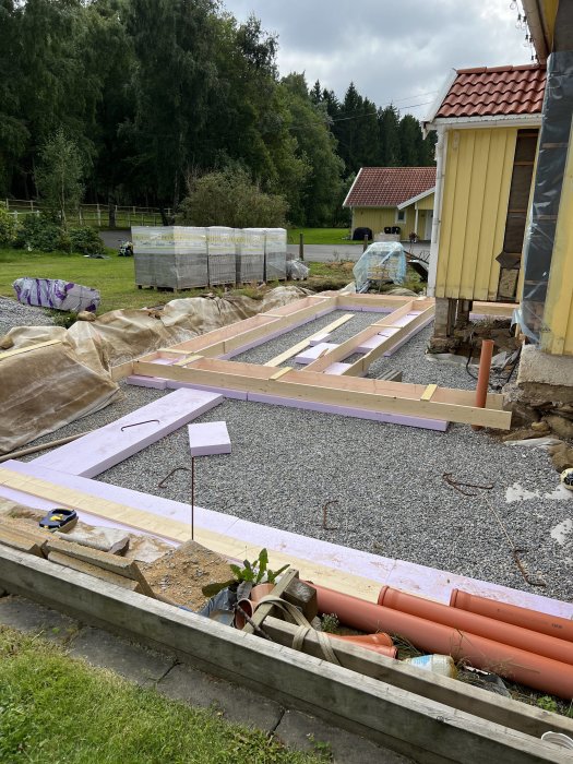 Grundarbeten för byggnation, träkonstruktioner, isoleringsmaterial, grus, husdel, byggmaterial, grönska, molnig himmel.