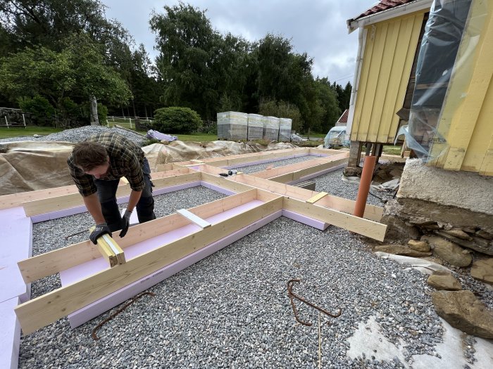 Person bygger trädäck utomhus nära hus, använder verktyg, trämaterial och isolering på grusunderlag.