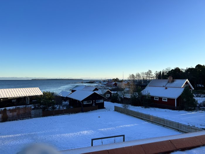 Vinterlandskap, snö, röda hus, klart himmel, havsutsikt, frodiga träd, soligt, lugnt bostadsområde.