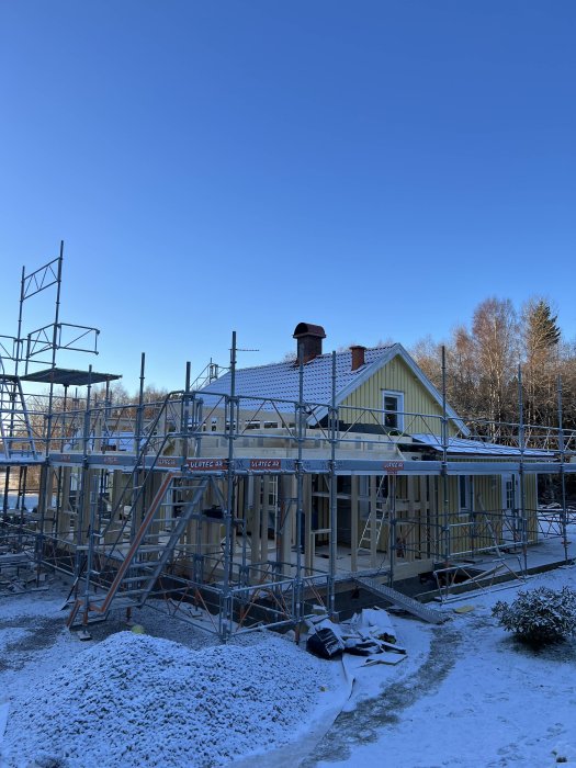 Husbyggnation under vinter, snö, ställning, klarblå himmel, träd bakgrunden, gul husfasad.