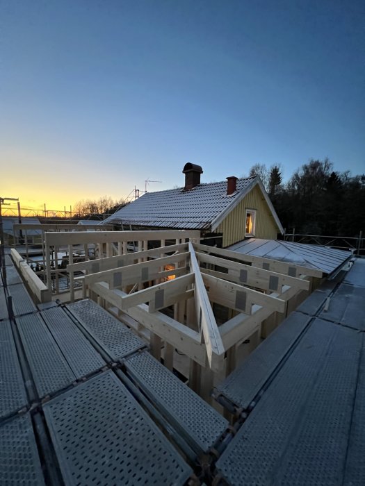 Trästomme för byggnation på tak vid solnedgång, med ett befintligt hus i bakgrunden.