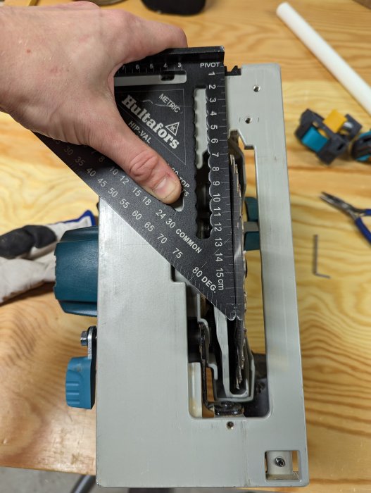 Hand håller vinkelhake mot geringssåg för att kontrollera sågvinkel. Träbearbetning, precision, verktyg, arbetsbänk synlig.