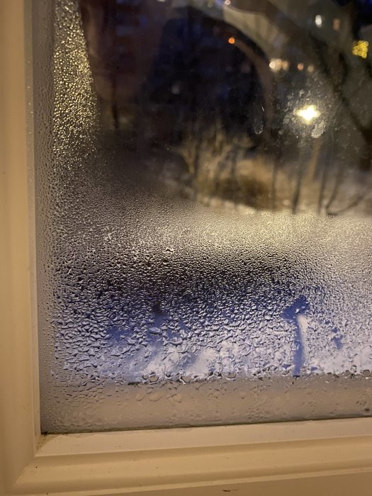 Fönster med kondens, suddig utsikt, natt, upplysta föremål i bakgrunden, vitt fönsterkarm.