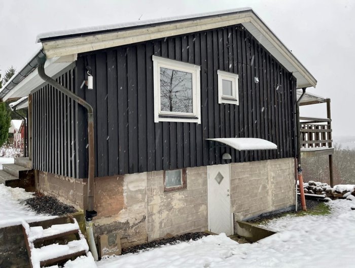 Svart trähus med snö, fönster, veranda, och delvis synlig murad grund. Snöfall och spår av grönska.