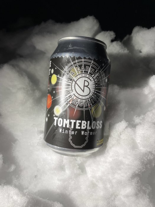 En ölburk märkt "Tomtebloss Winter Warmer", placerad på snö, med en mörk bakgrund och ljus belysning.