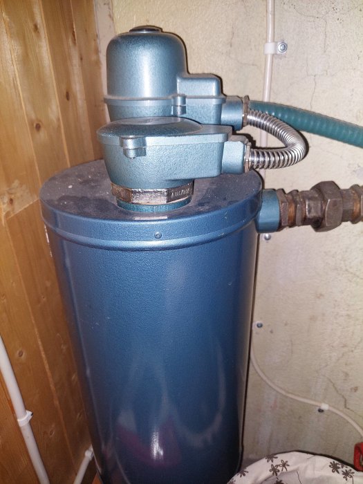 Blå varmvattenberedare med metallrör, installationsutrymme, träpanel och texturerad vägg i bakgrunden.