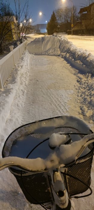 Snötäckt gångväg nattetid med cykelkorg i förgrunden och gatlyktor i bakgrunden.