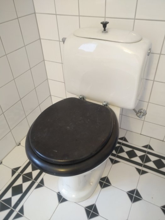 Toalett med svart sits, hög spolcistern, svartvit kakel, ren och traditionell design.