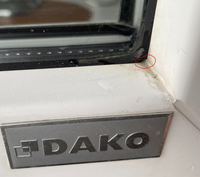 Fönster med DAKO-logotyp, mögel vid hörnet, röd cirkel markerar problemområde, vattendroppar på karmen.