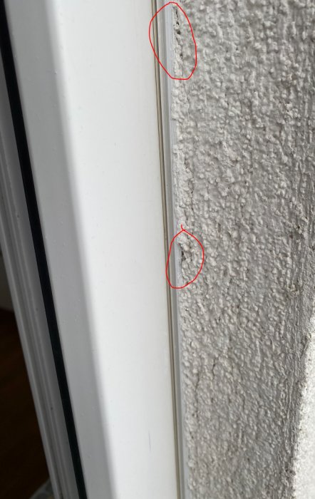 Skadad vägg intill fönsterkarm, hål i putsen, underliggande material synligt, behöver lagas.