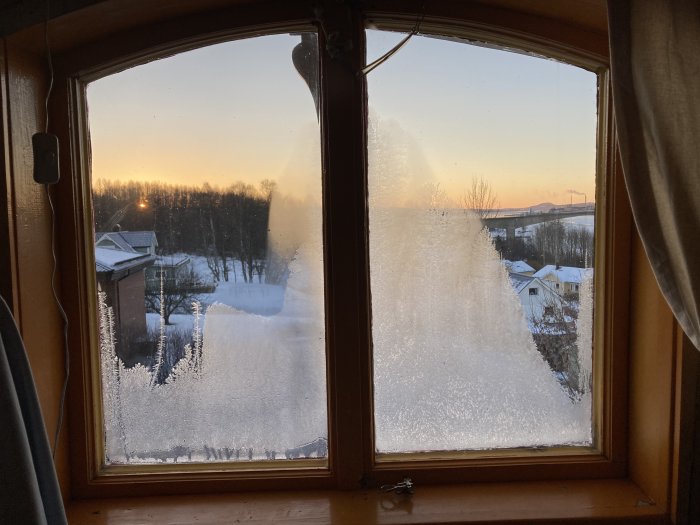 Solnedgång, vinterlandskap genom fönster med frost, varm inomhusutsikt, snötäckta tak, träd, avlägsen horisont.